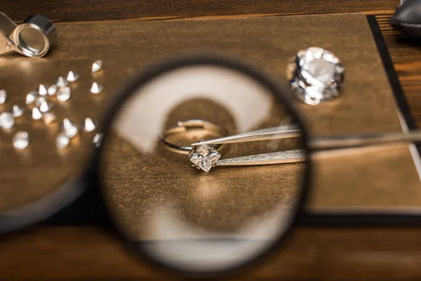 Selective focus of magnifying glass, jewelry ring with gemstone in tweezers on board on wooden table — Stock Photo