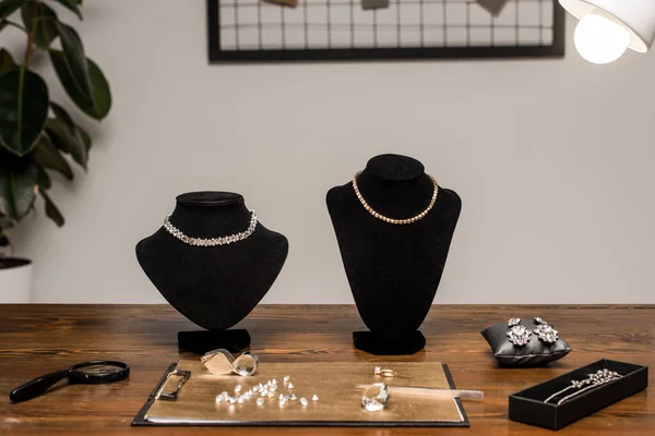 Jewelry necklaces on necklace stands near gemstones and magnifying glasses on board on table in workshop — Stock Photo