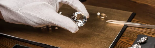 Vista recortada de la joyería tasador celebración de piedras preciosas cerca de joyas a bordo en la mesa de madera, tiro panorámico - foto de stock