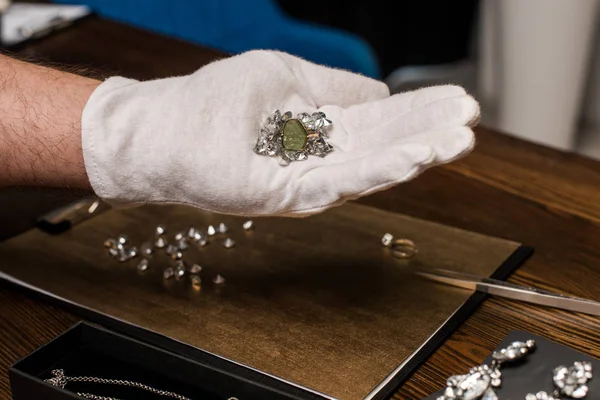 Обрезанный вид ювелира оценщик держа кольцо и драгоценные камни рядом ювелирные изделия и доска на столе — стоковое фото