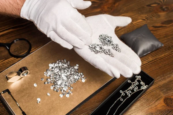 Cropped view of jewelry appraiser holding earrings near gemstones and magnifying glasses on board on table — Stock Photo