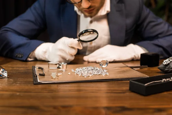 Vue recadrée de l'évaluateur de bijoux examinant les pierres précieuses à bord près des bijoux sur la table isolée sur noir — Photo de stock