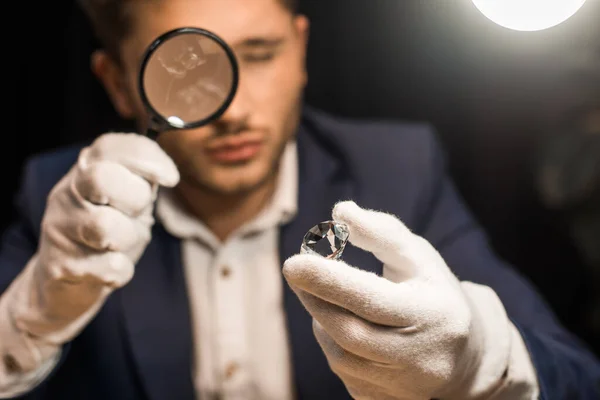 Selective focus of jewelry appraiser examining gemstone with magnifying glass on black background — Stock Photo