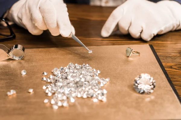 Ausgeschnittene Ansicht eines Schmuckgutachters, der Edelstein in einer Pinzette in der Nähe der Tafel auf dem Tisch hält — Stockfoto