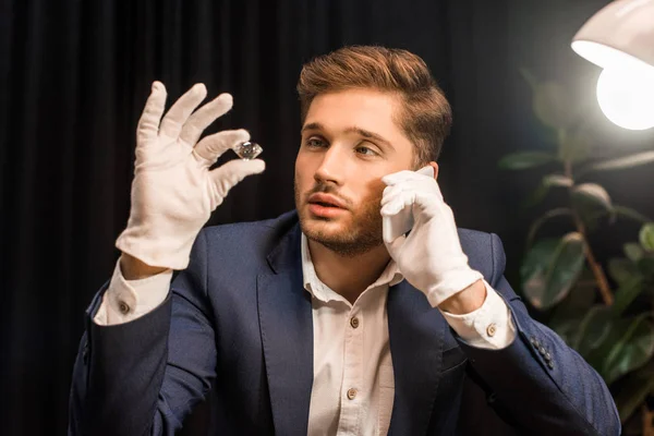 Evaluador de joyas hablando por teléfono inteligente y sosteniendo piedras preciosas en el taller - foto de stock