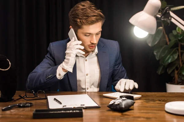 Avaliador de jóias falando no smartphone e usando calculadora perto de jóias na mesa — Fotografia de Stock