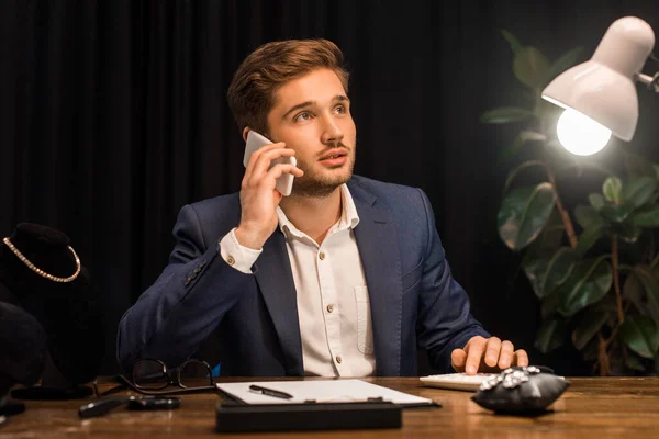 Pensativo tasador de joyas hablando en el teléfono inteligente y el uso de la calculadora cerca de joyas en la mesa en el taller - foto de stock