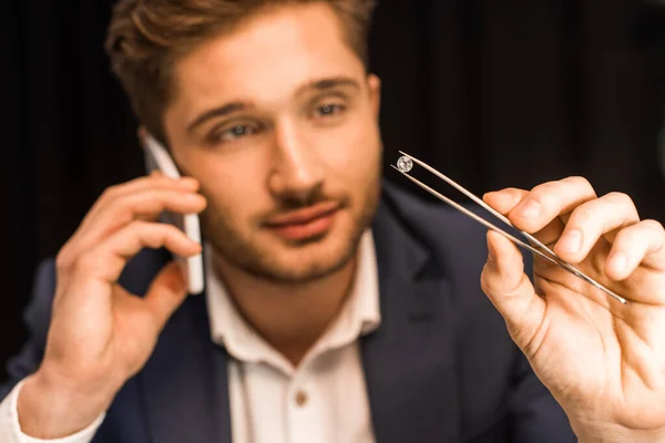 Selective focus of jewelry appraiser talking on smartphone and holding gemstone in tweezers isolated on black — Stock Photo