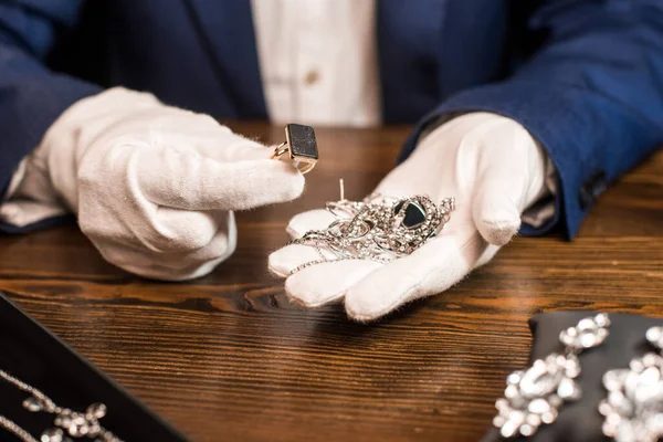 Vue recadrée de l'évaluateur de bijoux tenant bague et bijoux à table isolé sur noir — Photo de stock