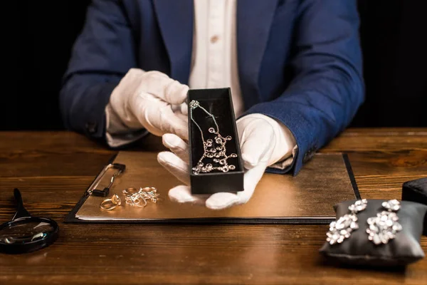 Cropped view of jewelry appraiser holding box with necklace near jewelry on board on table isolated on black — Stock Photo