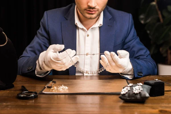 Vue recadrée de l'évaluateur de bijoux tenant collier près de bijoux à bord sur la table dans l'atelier — Photo de stock