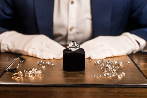 Vista recortada de piedras preciosas cerca de joyas a bordo y tasador de joyas en la mesa aislado en negro - foto de stock