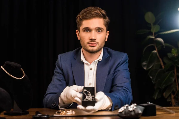 Jewelry appraiser looking at camera and holding necklace near jewelry on table in workshop — Stock Photo