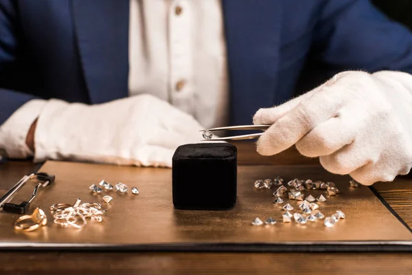 Vista recortada de la joyería tasador celebración de piedras preciosas en pinzas cerca de joyas a bordo de la mesa aislado en negro - foto de stock