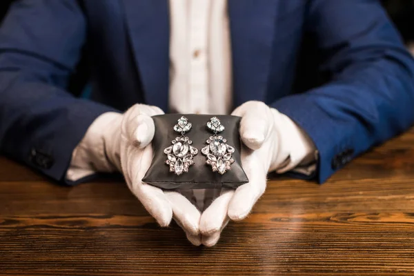 Vista cortada do avaliador de jóias segurando travesseiro de jóias com brincos na mesa isolada em preto — Fotografia de Stock