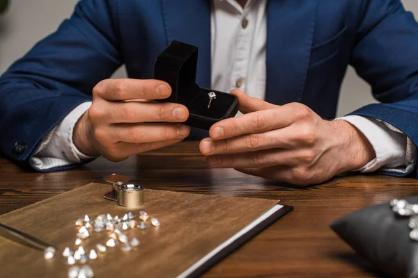 Vue recadrée de l'évaluateur de bijoux tenant boîte avec bague de bijoux près des pierres précieuses sur la table sur fond gris — Photo de stock