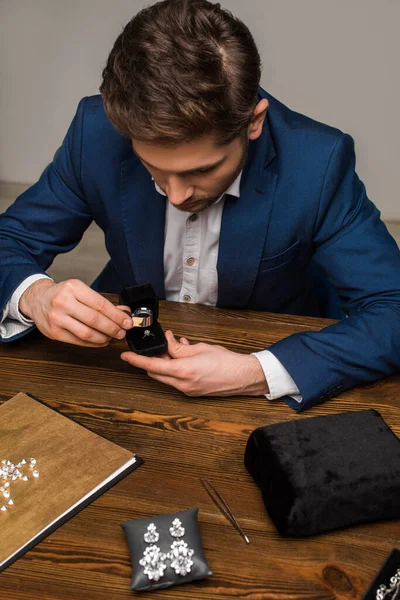 Schmuckgutachter untersucht Schmuckring mit Lupe in der Nähe von Schmuck auf Tisch in Werkstatt — Stockfoto