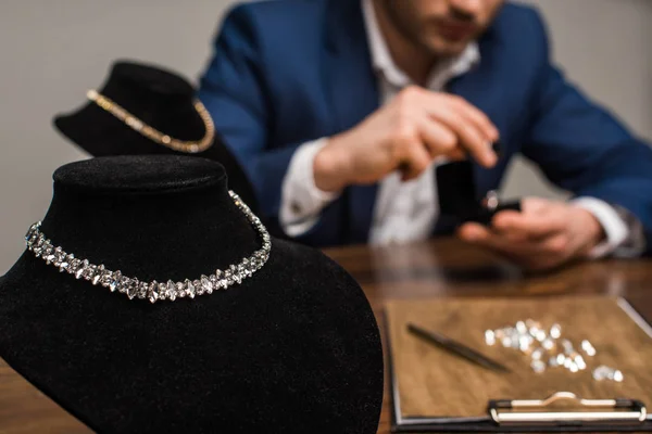 Селективный фокус ожерелий на стендах ожерелий и ювелирный оценщик, работающий за столом, изолированным на сером — стоковое фото