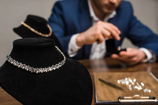 Selective focus of necklaces on necklace stands and jewelry appraiser working at table isolated on grey — Stock Photo