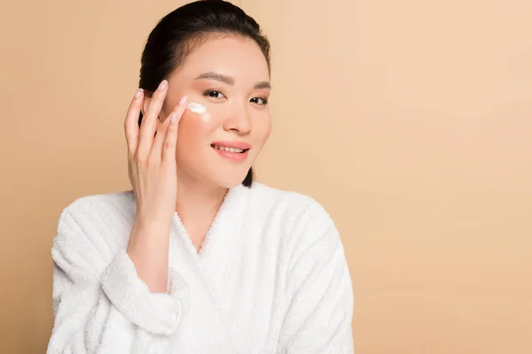 Sonriente hermosa mujer asiática en albornoz aplicando crema facial sobre fondo beige - foto de stock