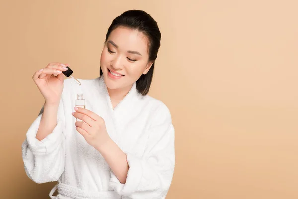 Sourire belle asiatique femme en peignoir tenant compte compte compte-gouttes et bouteille avec sérum sur fond beige — Photo de stock