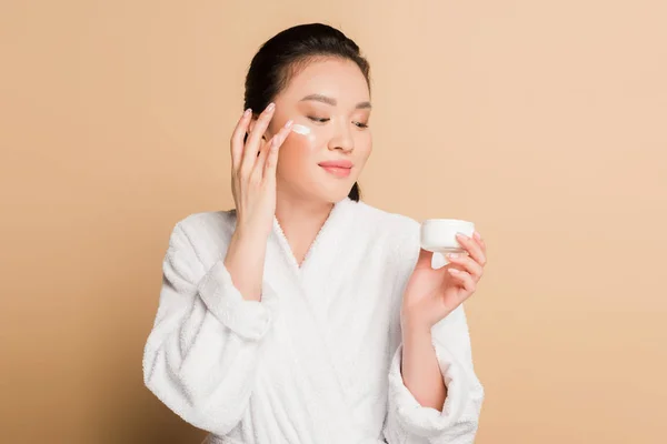 Sorrindo bela mulher asiática em roupão aplicando creme facial no fundo bege — Fotografia de Stock