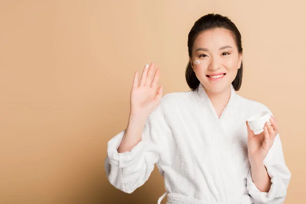 Sorridente bella donna asiatica in accappatoio con crema viso sulla guancia e dito su sfondo beige — Foto stock