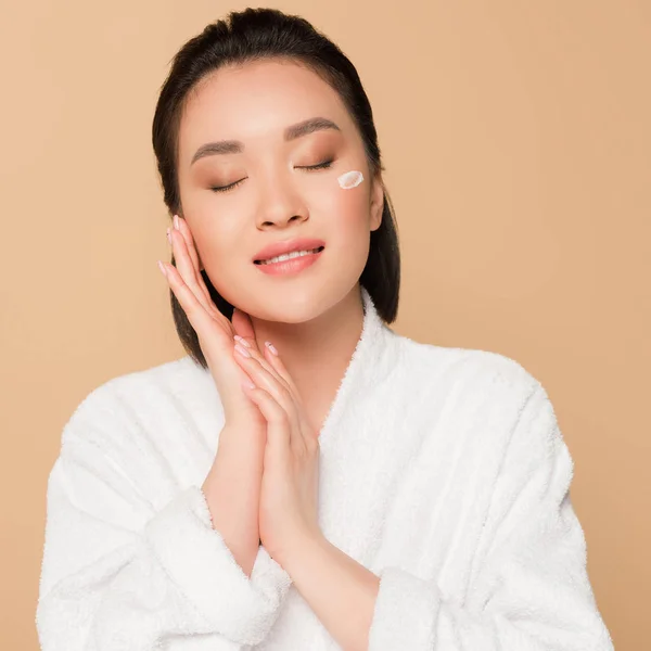 Pleased beautiful asian woman in bathrobe with face cream on cheek and closed eyes on beige background — Stock Photo