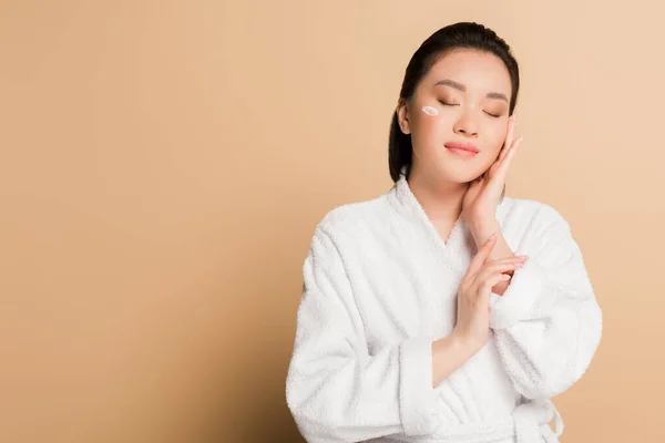 Pleased beautiful asian woman in bathrobe with face cream on cheek and closed eyes on beige background — Stock Photo