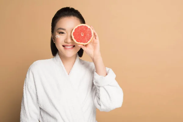 Smiling beautiful asian woman in bathrobe with face cream on cheek and grapefruit half on beige background — Stock Photo