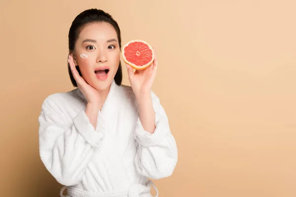 Shocked beautiful asian woman in bathrobe with face cream on cheek and grapefruit half on beige background — Stock Photo
