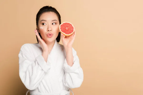 Surprised beautiful asian woman in bathrobe with face cream on cheek and grapefruit half on beige background — Stock Photo