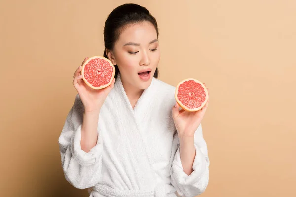 Shocked beautiful asian woman in bathrobe with grapefruit halves on beige background — Stock Photo