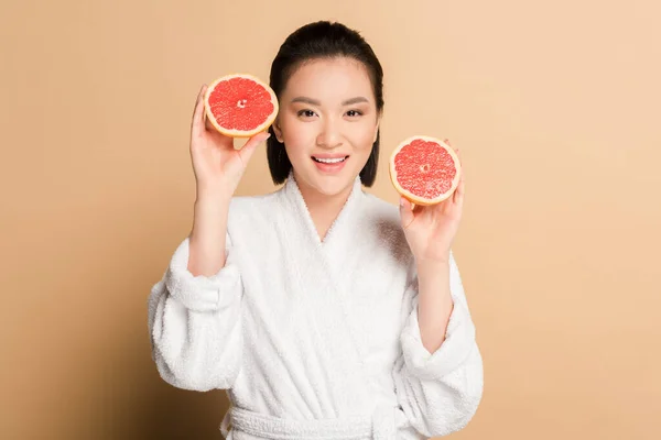 Glücklich schöne asiatische Frau im Bademantel mit Grapefruithälften auf beigem Hintergrund — Stockfoto