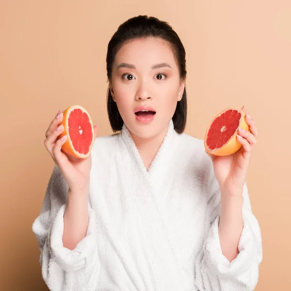 Shocked beautiful asian woman in bathrobe with grapefruit halves on beige background — Stock Photo