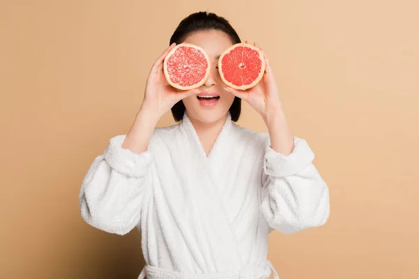 Impactada hermosa mujer asiática en albornoz con mitades de pomelo en los ojos sobre fondo beige - foto de stock