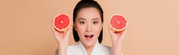 Impactado hermosa mujer asiática en albornoz con mitades de pomelo aislado en beige, tiro panorámico — Stock Photo