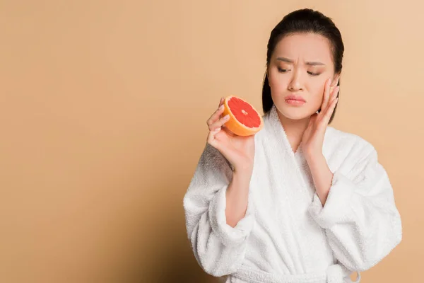 Сумна красива азіатська жінка в халаті з половинками грейпфрута на бежевому фоні — стокове фото