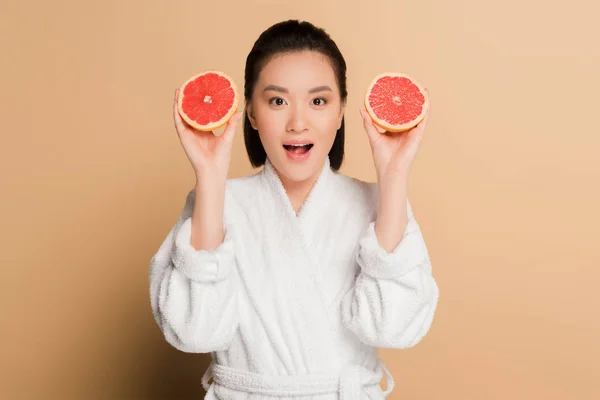 Impactada hermosa mujer asiática en albornoz con mitades de pomelo sobre fondo beige - foto de stock