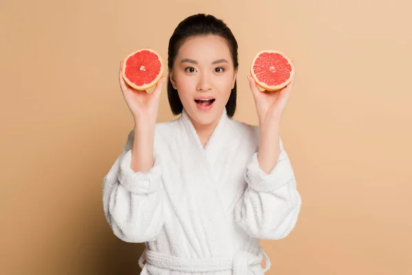 Shocked beautiful asian woman in bathrobe with grapefruit halves on beige background — Stock Photo