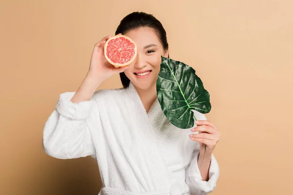 Glücklich schöne asiatische Frau im Bademantel mit Grapefruit-Hälfte und Blatt auf beigem Hintergrund — Stockfoto