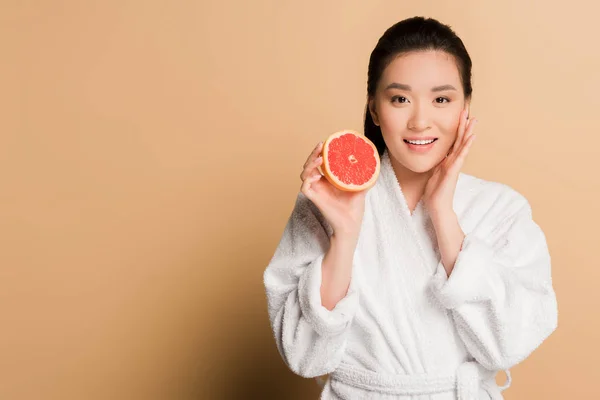 Happy beautiful asian woman in bathrobe with grapefruit half on beige background — Stock Photo