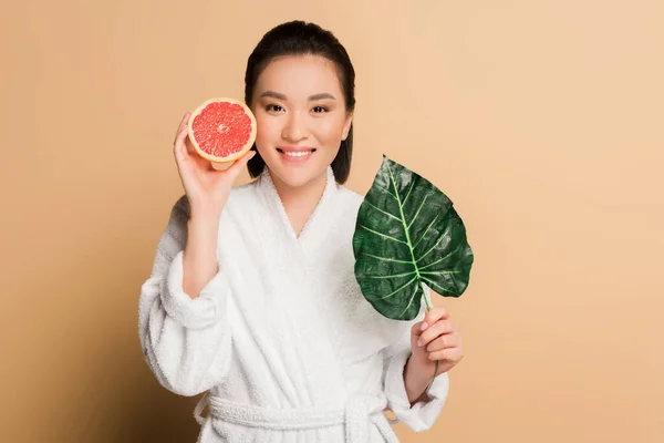 Щаслива красива азіатська жінка в халаті з грейпфрутом наполовину і листком на бежевому фоні — стокове фото