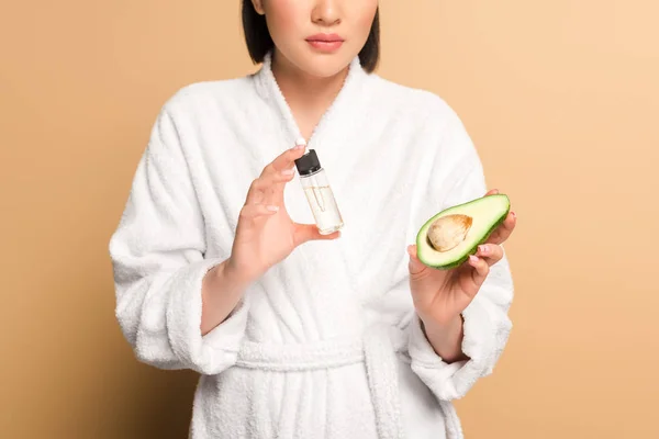 Cropped view of beautiful asian woman in bathrobe with avocado half and oil in bottle on beige background — Stock Photo