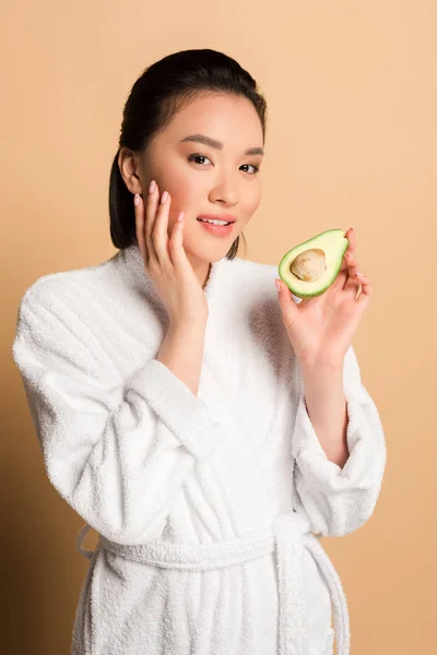 Beautiful asian woman in bathrobe with avocado half touching face on beige background — Stock Photo