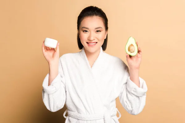 Happy beautiful asian woman in bathrobe with avocado half and face cream on beige background — Stock Photo