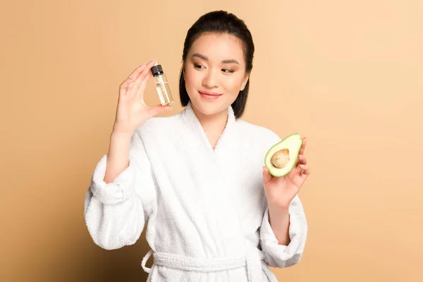 Glücklich schöne asiatische Frau im Bademantel mit Avocado-Hälfte und Öl in der Flasche auf beigem Hintergrund — Stockfoto