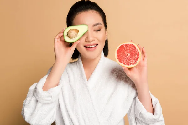 Happy beautiful asian woman in bathrobe with avocado and grapefruit halves on beige background — Stock Photo