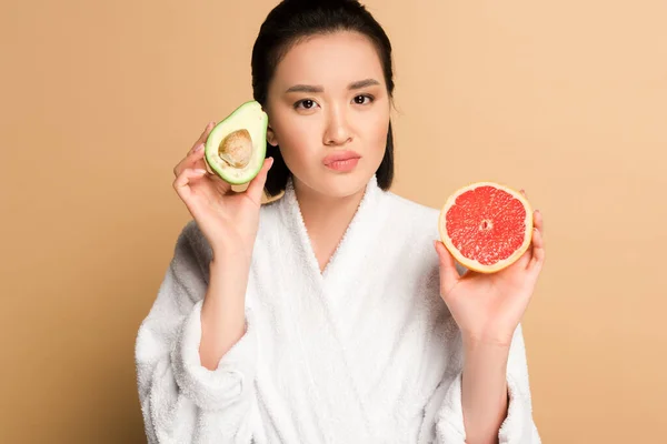 Skeptical beautiful asian woman in bathrobe with avocado and grapefruit halves on beige background — Stock Photo