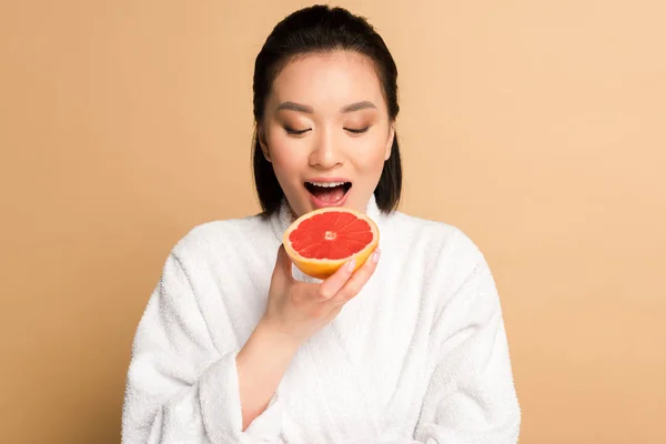 Beautiful asian woman in bathrobe eating grapefruit on beige background — Stock Photo